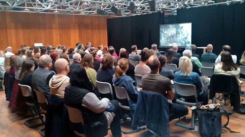 Blick in die Runde der Teilnehmenden; Foto zeigt sitzende Teilnehmende an der Veranstaltung mit Blick in Richtung Redner, der an einem Pult steht. Im Hintergrund ist eine Präsentationswand zu sehen.