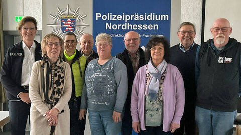 Austauschtreffen der SIcherheitsberatung für Seniorinnen und Senioren bei der Polizeidirektion Waldeck-Frankenberg; Mehrere Personen stehen vor einer Leinwand mit dem Polizeistern und dem Logo des Polizeipräsidiums Nordhessen