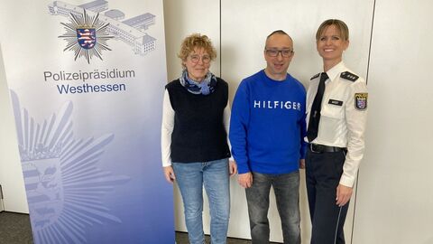 Frau Nickel, Herr Hippler und Polizeihauptkommissarin Wüst vor dem Logo des Polizeipräsidiums Westhessen stehend